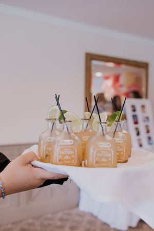 A hand holds a tray of mason jar cocktails garnished with lime and black straws on white linens.