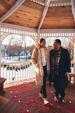 A couple stands in a gazebo with scattered rose petals on the wooden floor during a winter sunset.