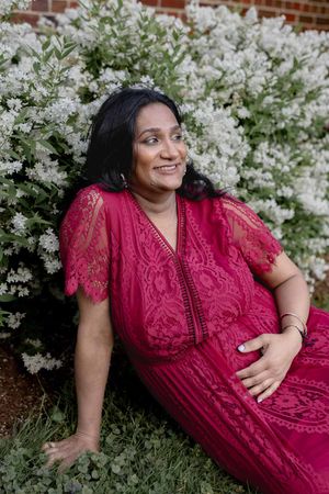 A smiling person in a red lace dress relaxes on grass near blooming white flowers and a brick wall.