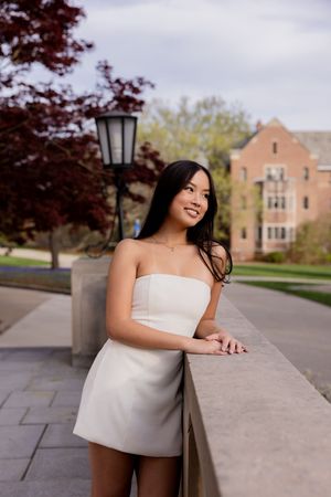 Figure leaning on stone wall wearing strapless white dress posing outdoors near brick building and trees.