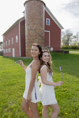Two friends in white dresses pose with bubble wands on a lawn in front of a classic red barn with a silo.