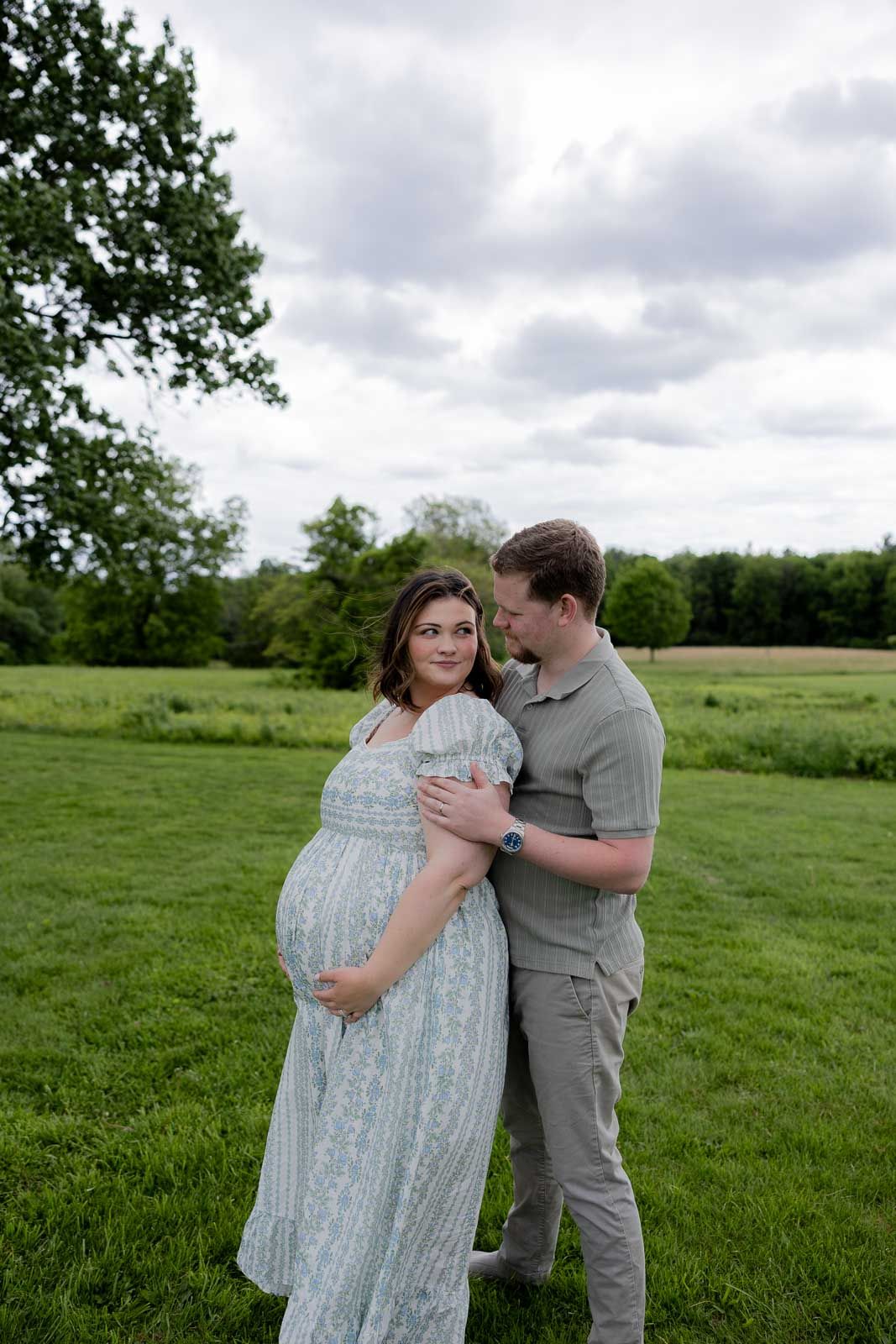 A couple embraces in a serene meadow setting, with the woman wearing a flowing lace dress against cloudy skies.