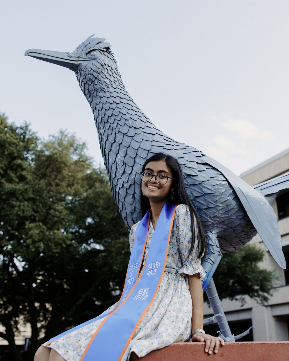 San Antonio Graduation. A graduate poses next to a large bird sculpture wearing graduation regalia and floral dress on a university campus.