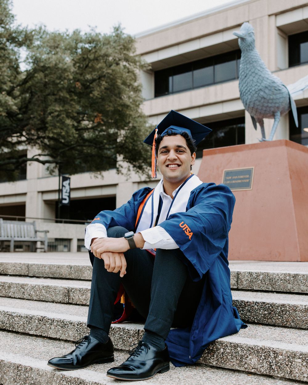 San Antonio Graduation. Graduate in blue cap and gown sits on concrete steps with a large bird sculpture visible in the background.
