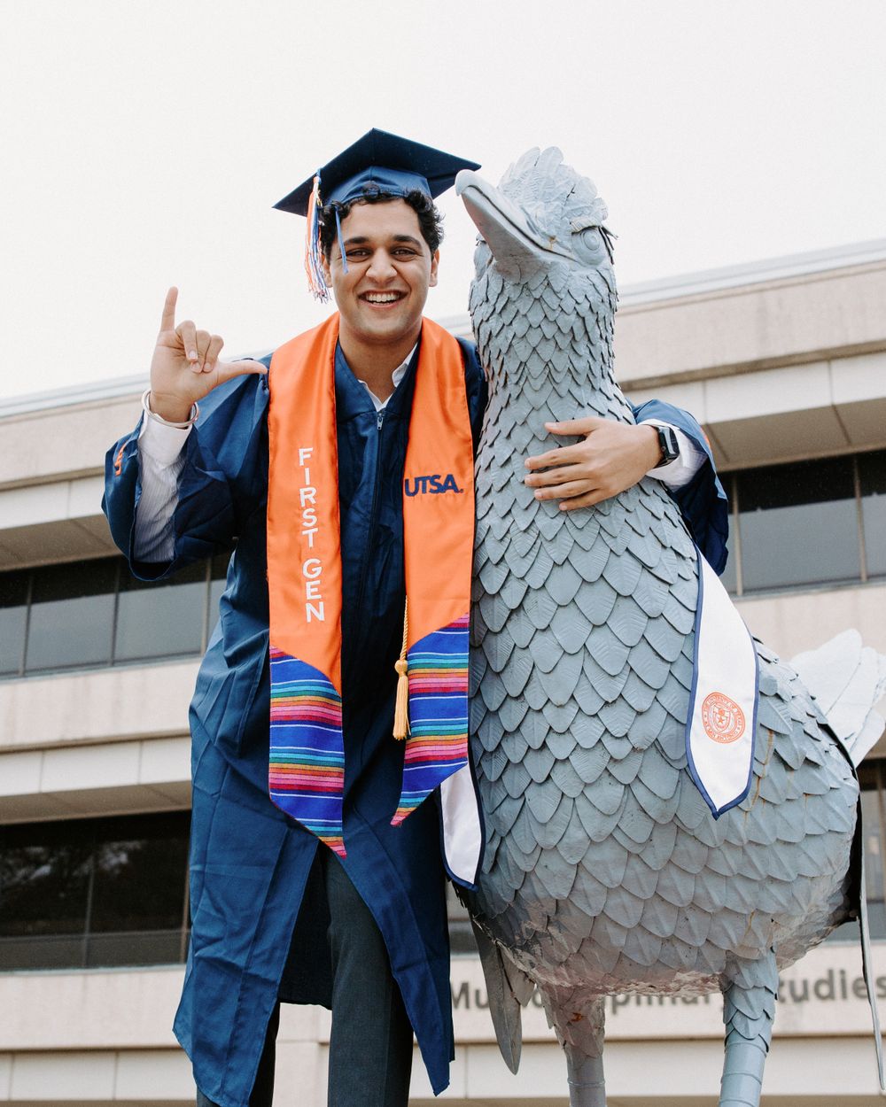 San Antonio Graduation. A graduate in cap and gown poses with a large bird statue while holding up one finger on a college campus.