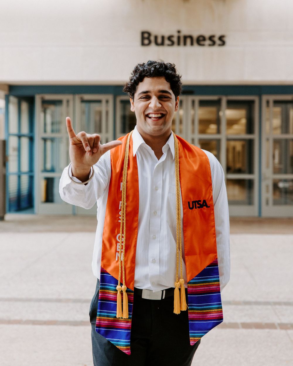 SA Graduate in white dress shirt and orange graduation stole with colorful stripes makes a celebratory gesture outside Business building.