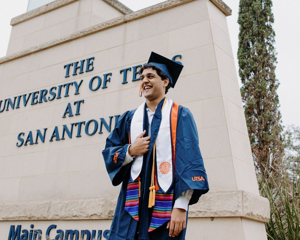 San Antonio Graduation. A graduate wearing academic regalia stands in front of The University of Texas at San Antonio main campus sign.