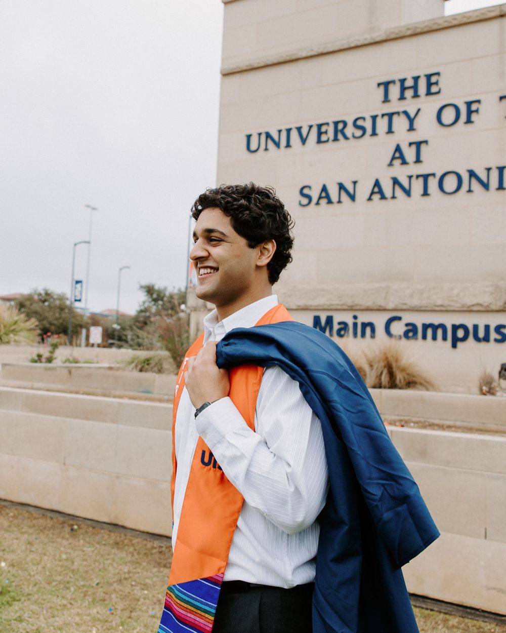 San Antonio Graduation. Student wearing orange and white attire stands smiling outside The University of San Antonio main campus building.