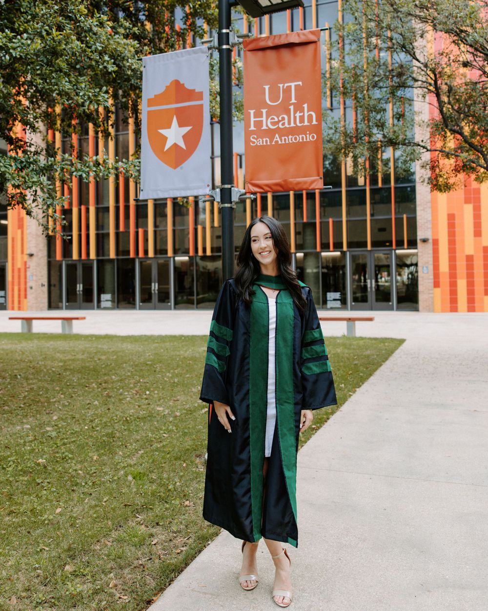 A graduate poses in traditional commencement regalia outside the UT Health San Antonio building with orange banners.