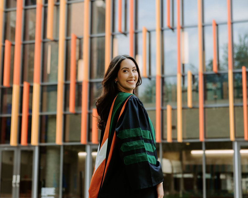 San Antonio Graduation. Person wearing a green stole turns while standing in front of a modern building with orange pillars.