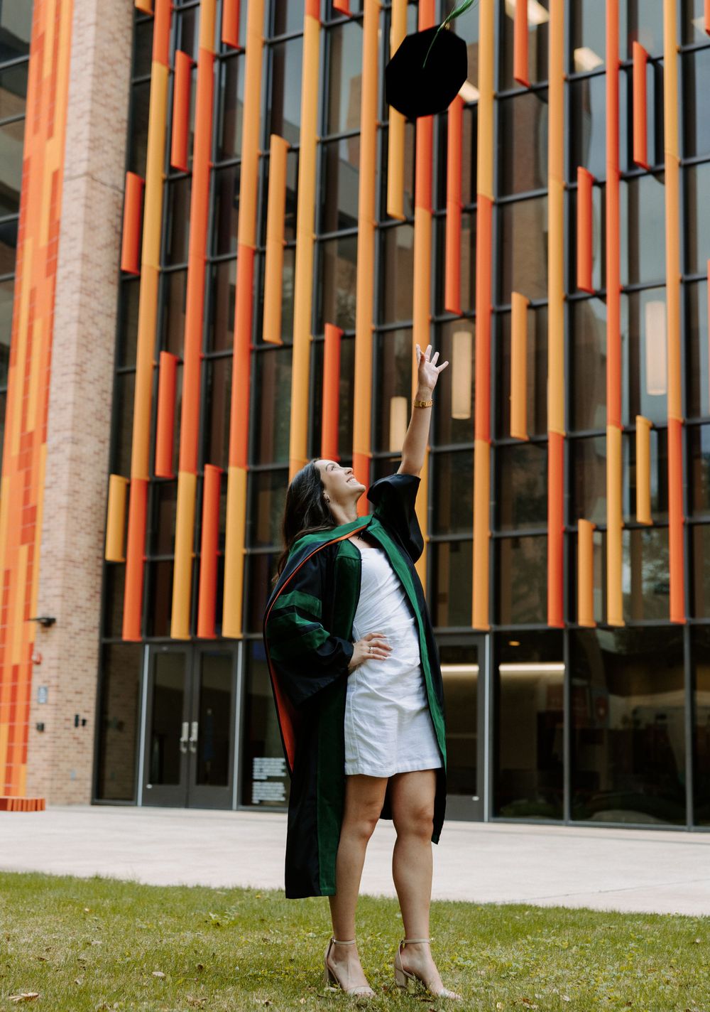 San Antonio Graduation. Graduate in cap and gown throwing graduation cap up in front of modern building with orange architectural details.