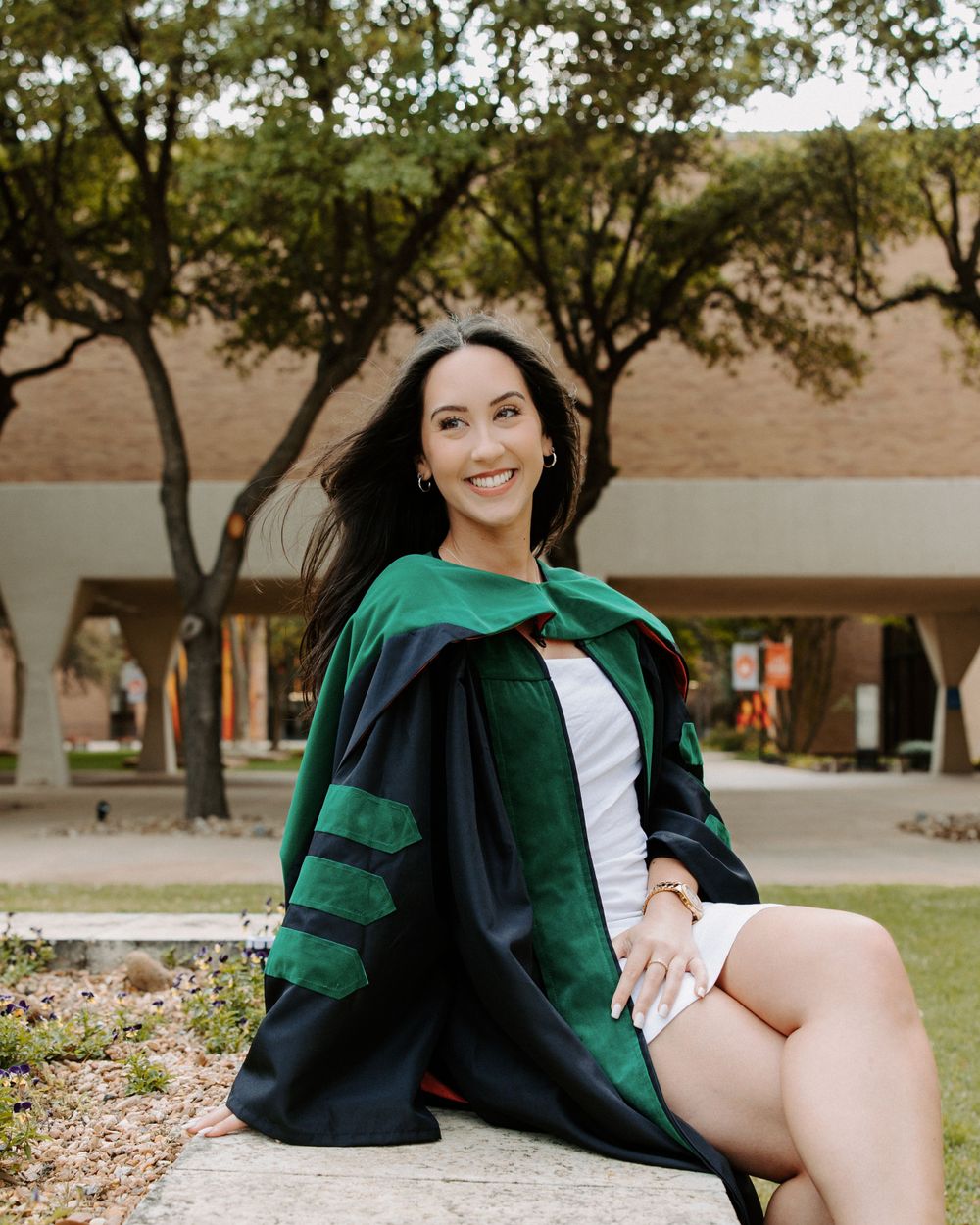 San Antonio Graduation. Graduate in green and black academic regalia sits smiling on a stone ledge in front of tree-lined academic building.