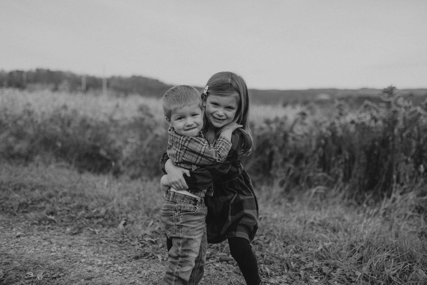 Two young children hugging and smiling together in a rural field setting.