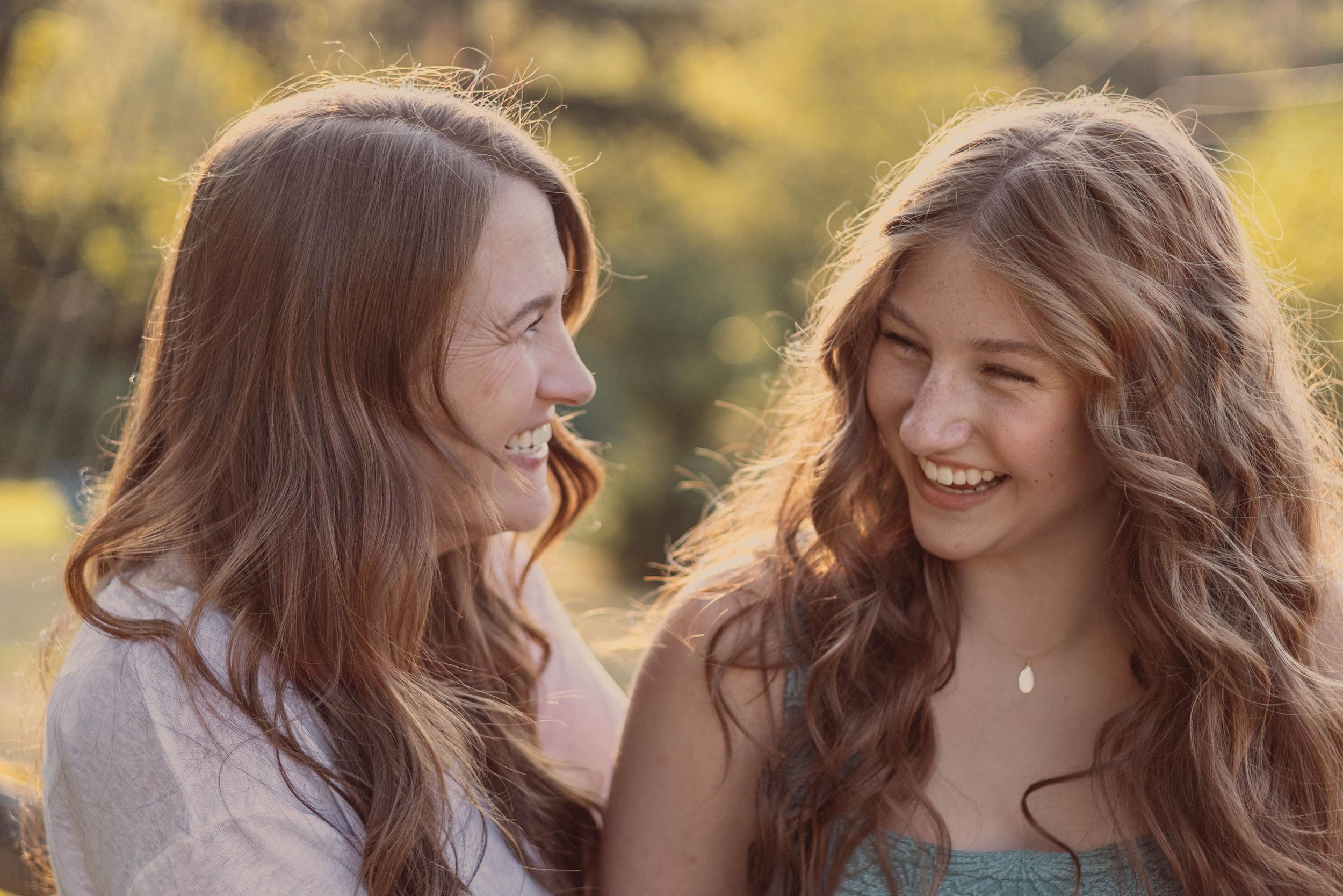 Two young women with long wavy hair sharing a candid laughing moment together in warm golden sunlight.