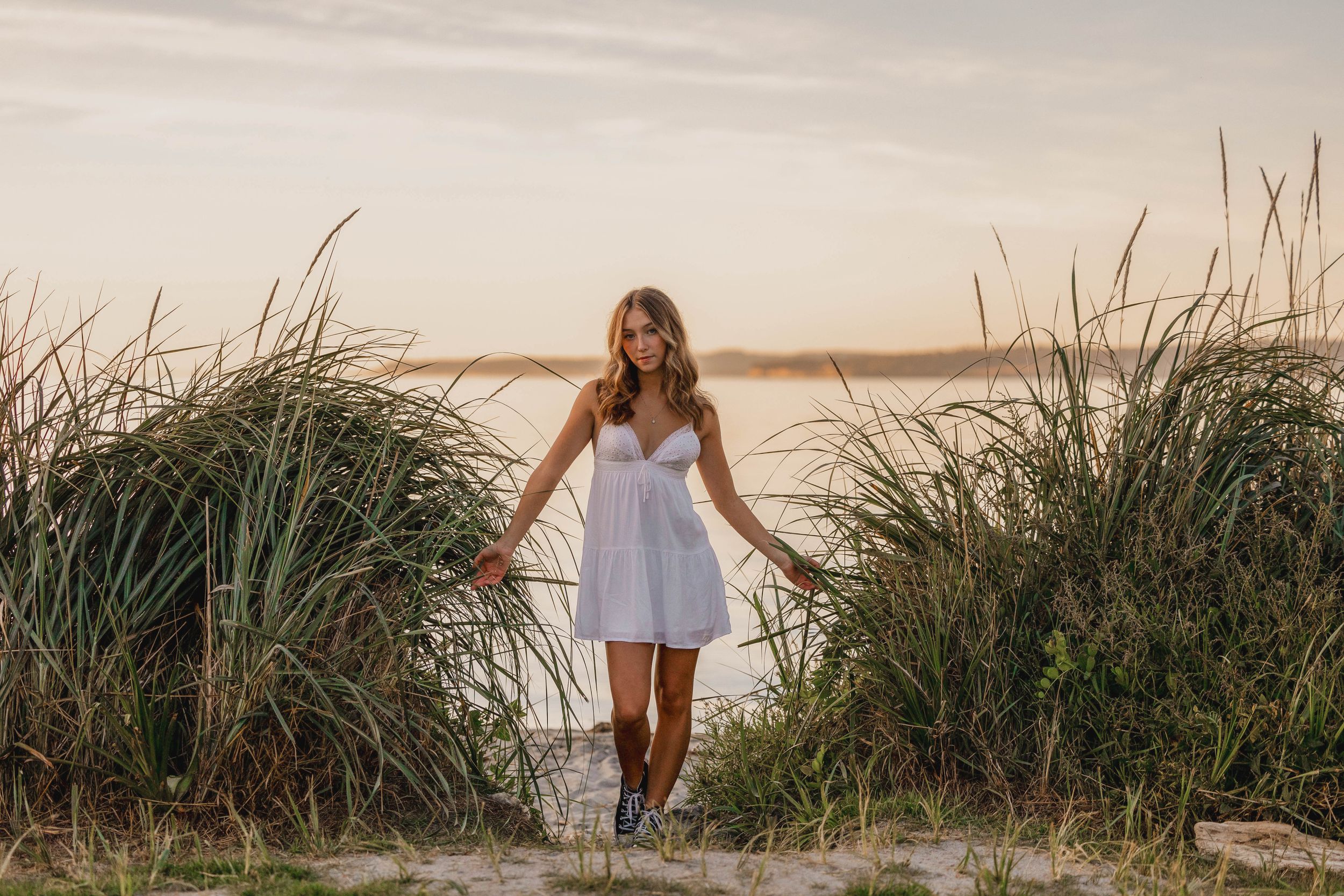 Woman in white dress walks through tall grass at sunset near water.