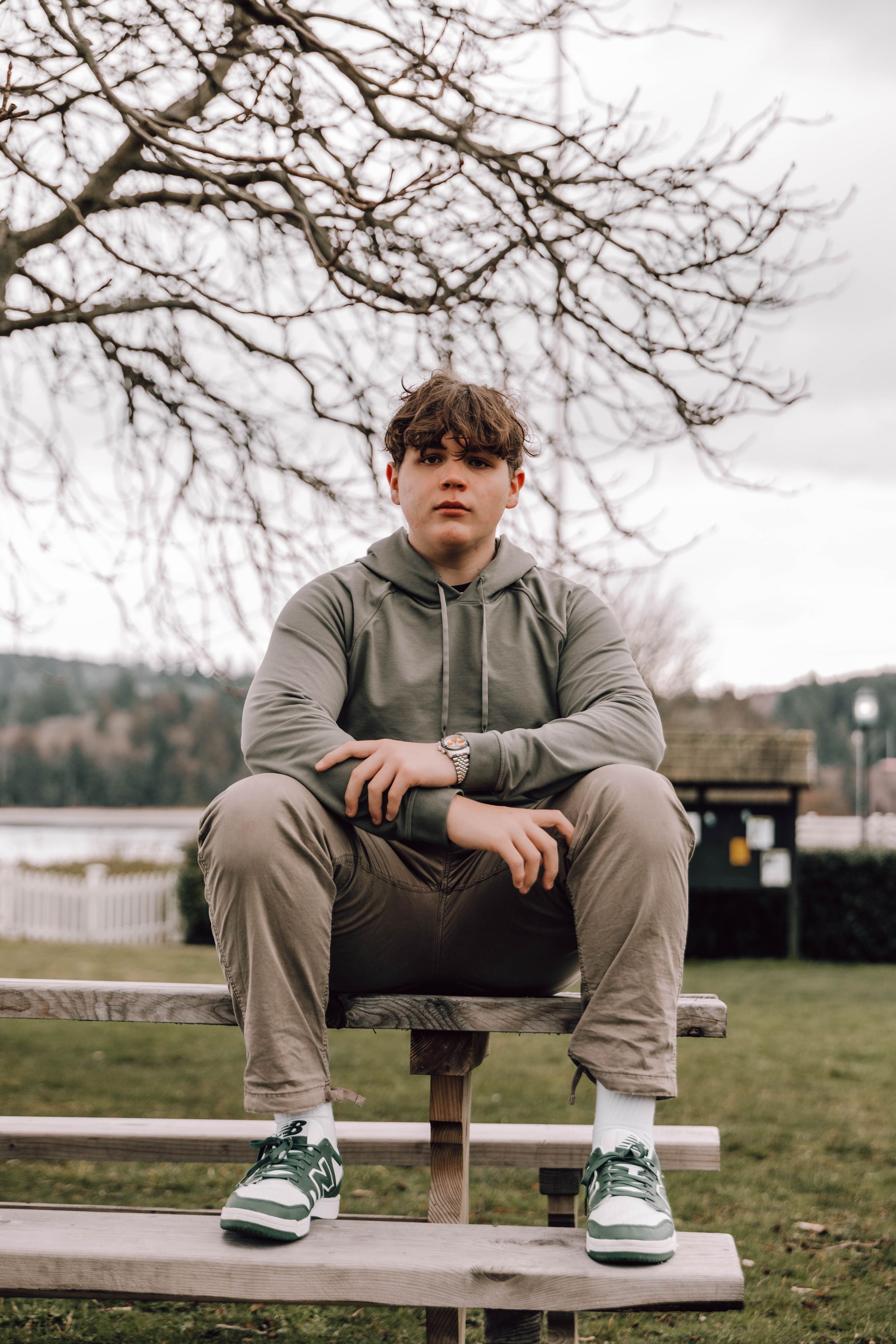 Person wearing a grey hoodie and casual pants sits outdoors on a bench wearing green sneakers in an overcast setting.