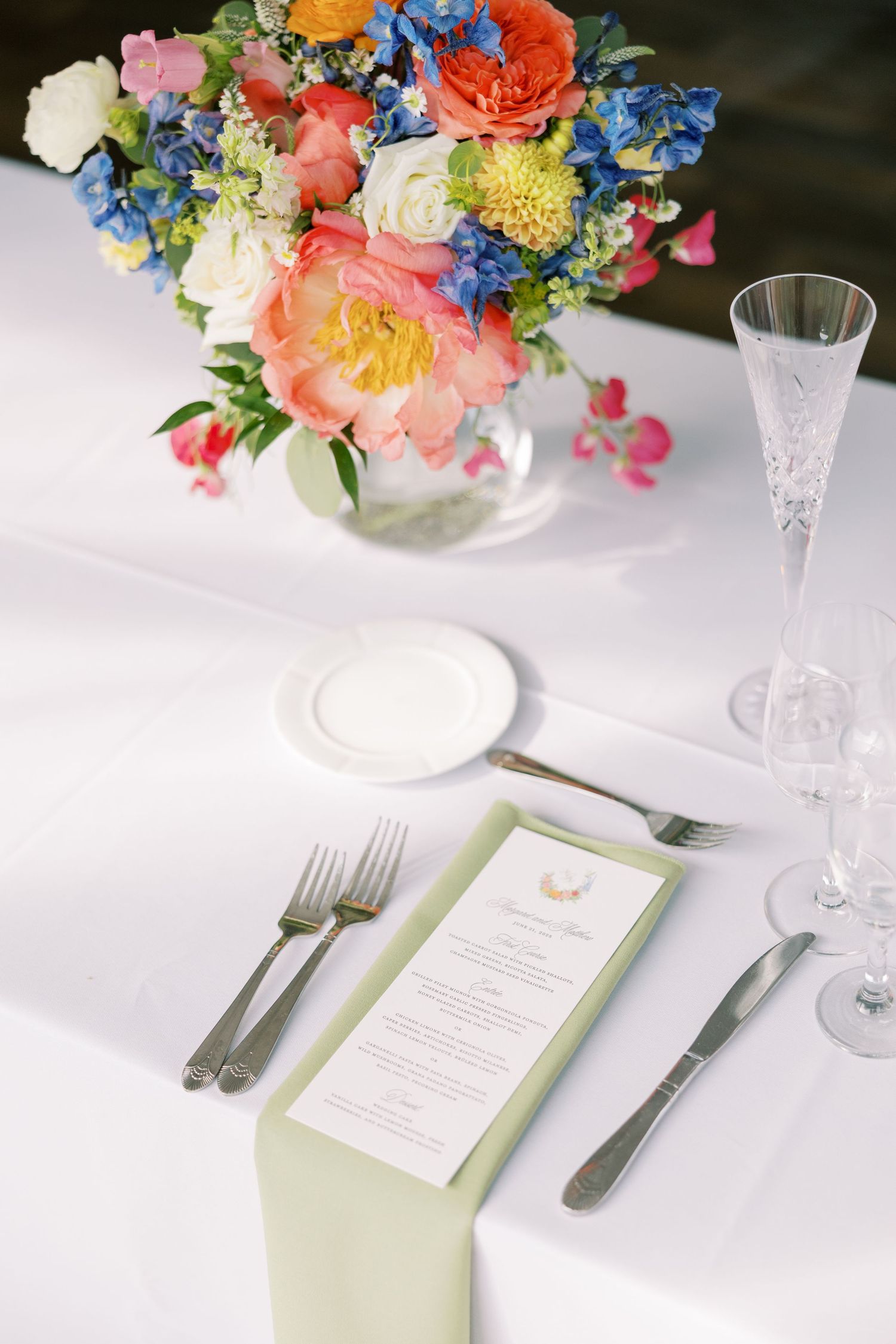 Elegant table setting with colorful floral centerpieces, white linens, crystal glasses, and menu cards with sage green accents.