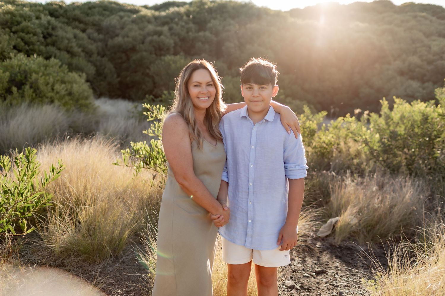 Two people standing together in natural sunlight on a path with dried grass and greenery in the background at sunset.