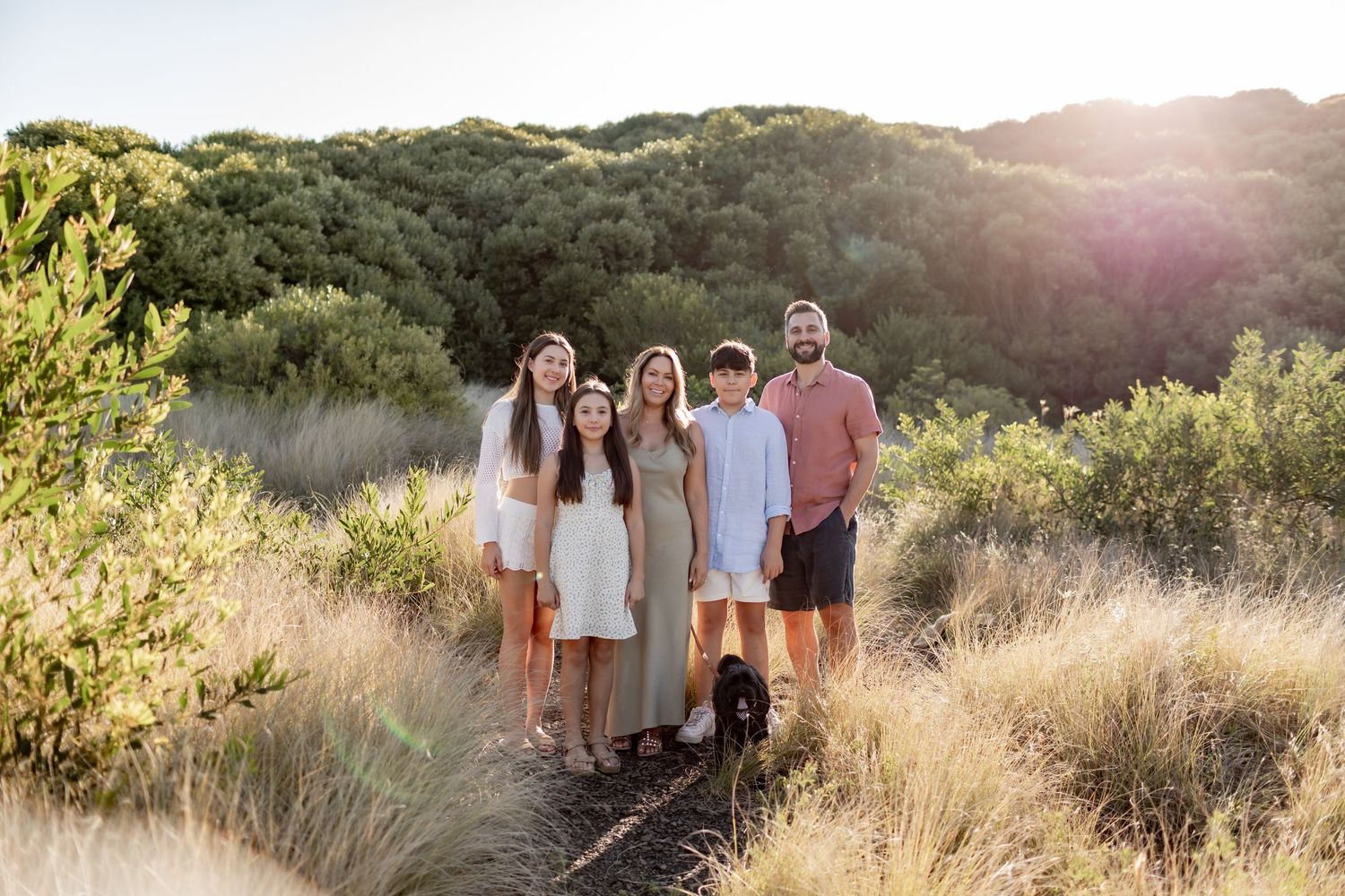 A group in light-colored clothing poses for outdoor family portraits in a scenic golden field at sunset.