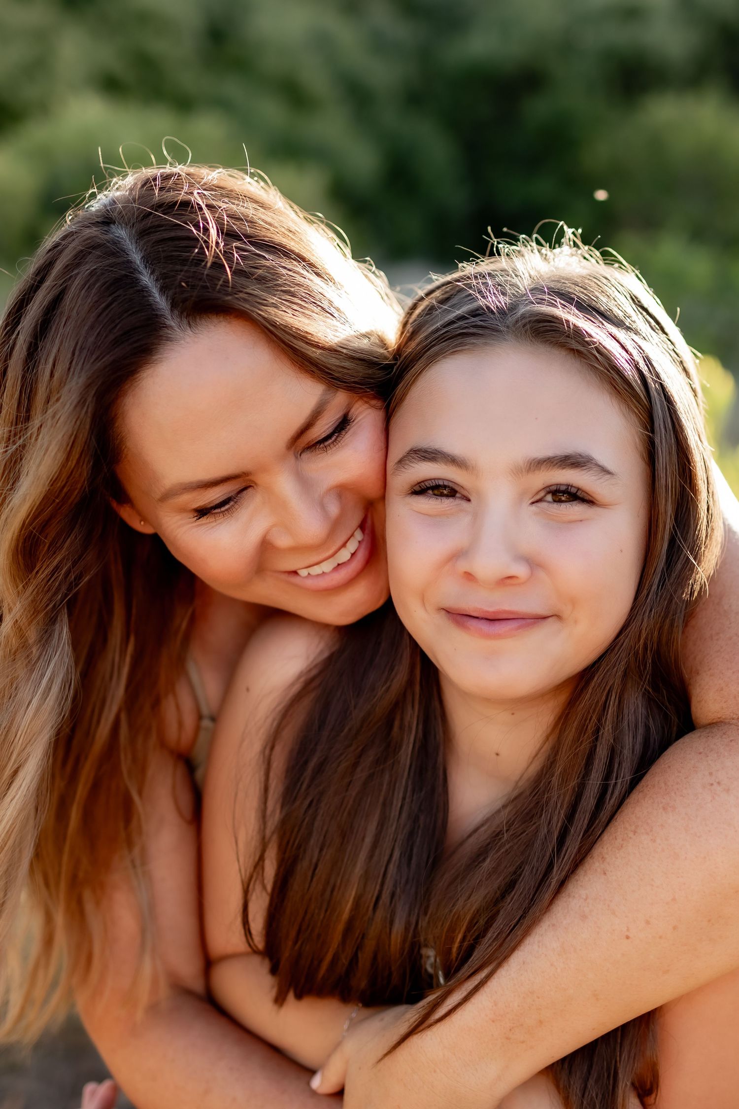 Two friends sharing a tender embrace and smiling while backlit by warm sunlight in an outdoor setting.