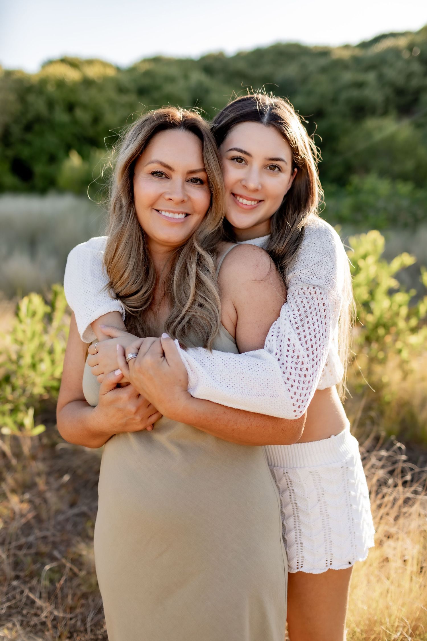 Two women embrace in a sun-filled outdoor setting wearing white and beige outfits.