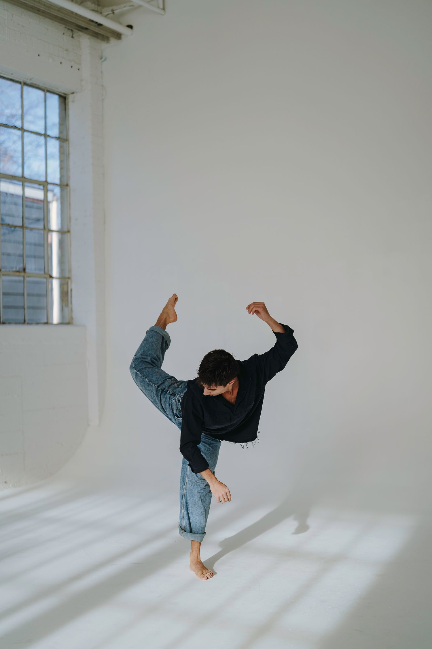 Person in denim jacket performs a dynamic dance move in a minimalist white studio with natural window light.