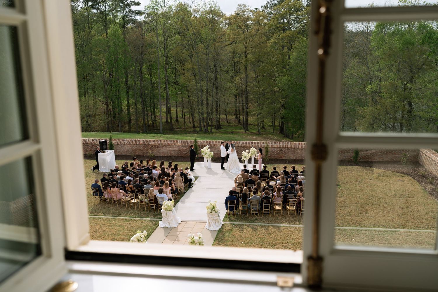Outdoor wedding ceremony viewed from above through an open window with wooded backdrop.