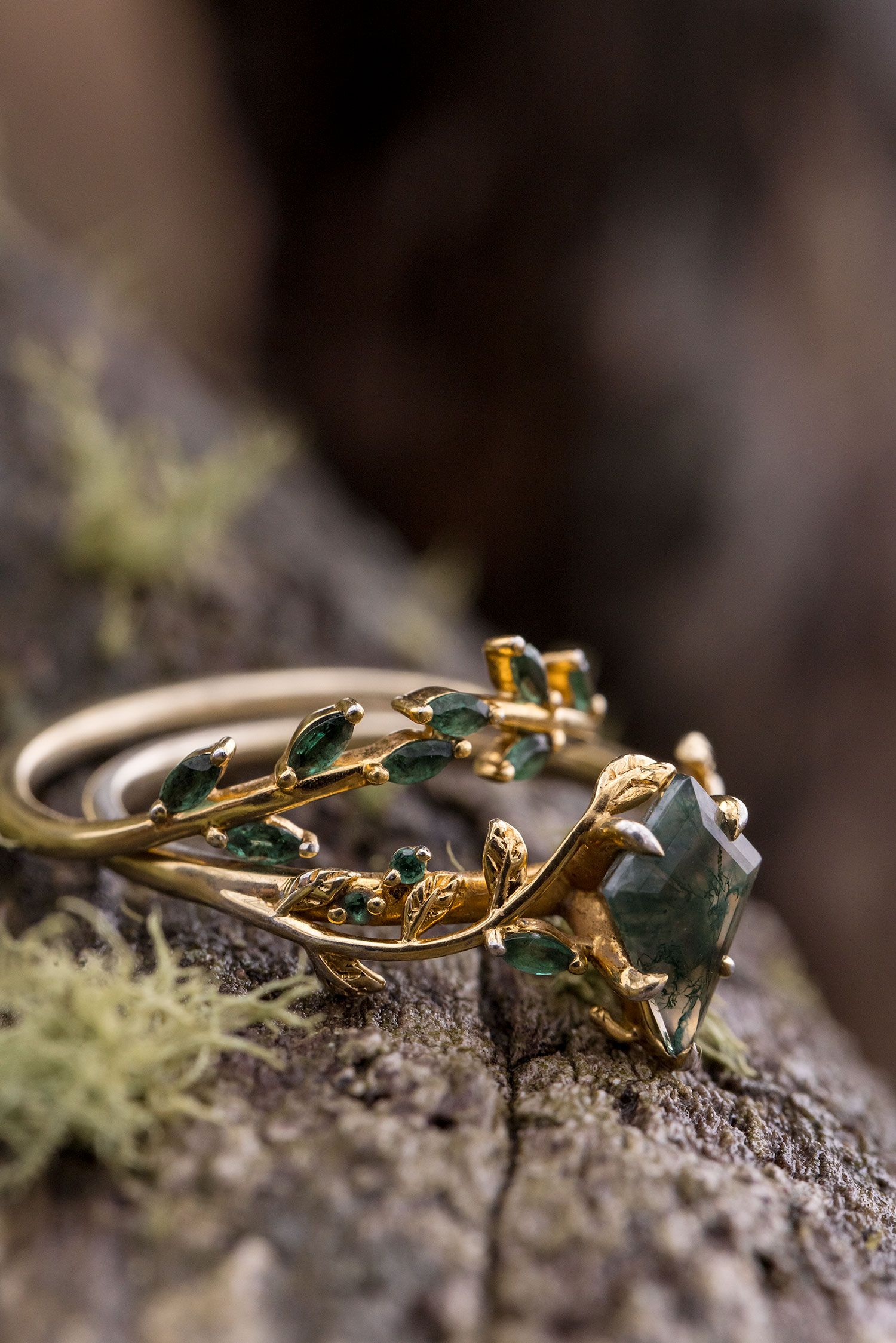 Delicate vintage-style gold ring with emerald gemstone and leaf details rests on mossy stone in macro photography.