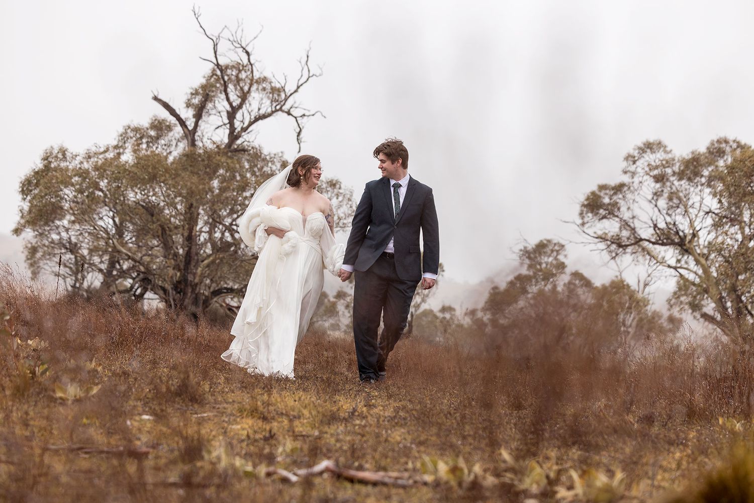 A wedding couple walks hand in hand through a misty Winter hillside with bare trees in the background in Jindabyne.
