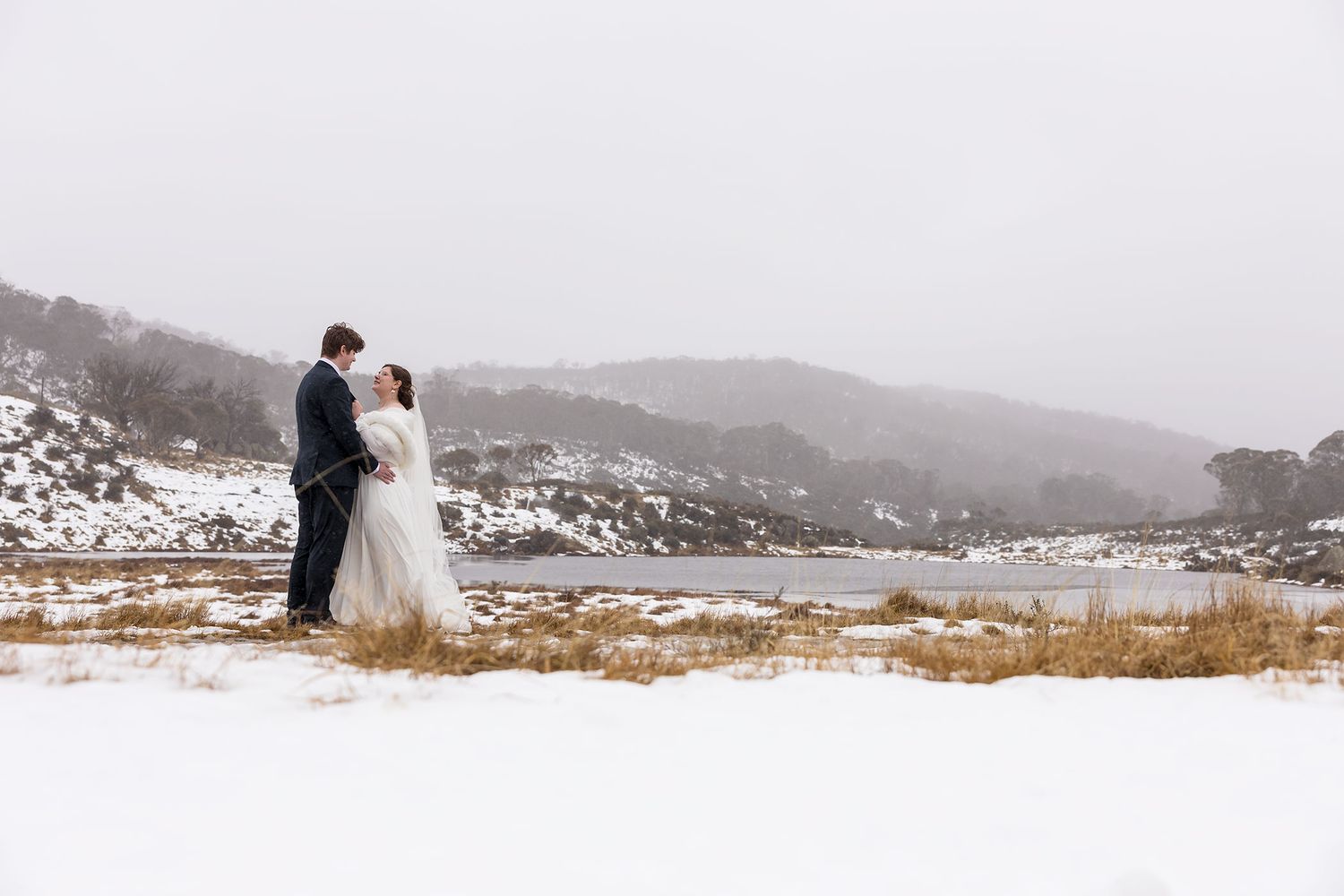 A winter wedding couple embraces on a snowy hilltop overlooking a serene mountainous landscape at Mount Perisher near Lake Jindabyne.