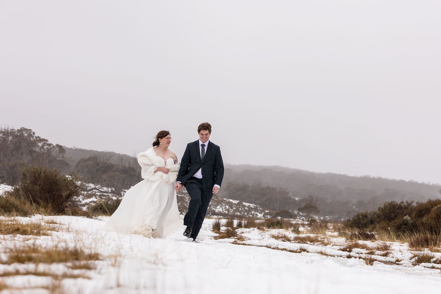 A couple in formal wedding attire laughs together while walking through fresh snow in a winter landscape at Mount Perisher near Jindabyne.