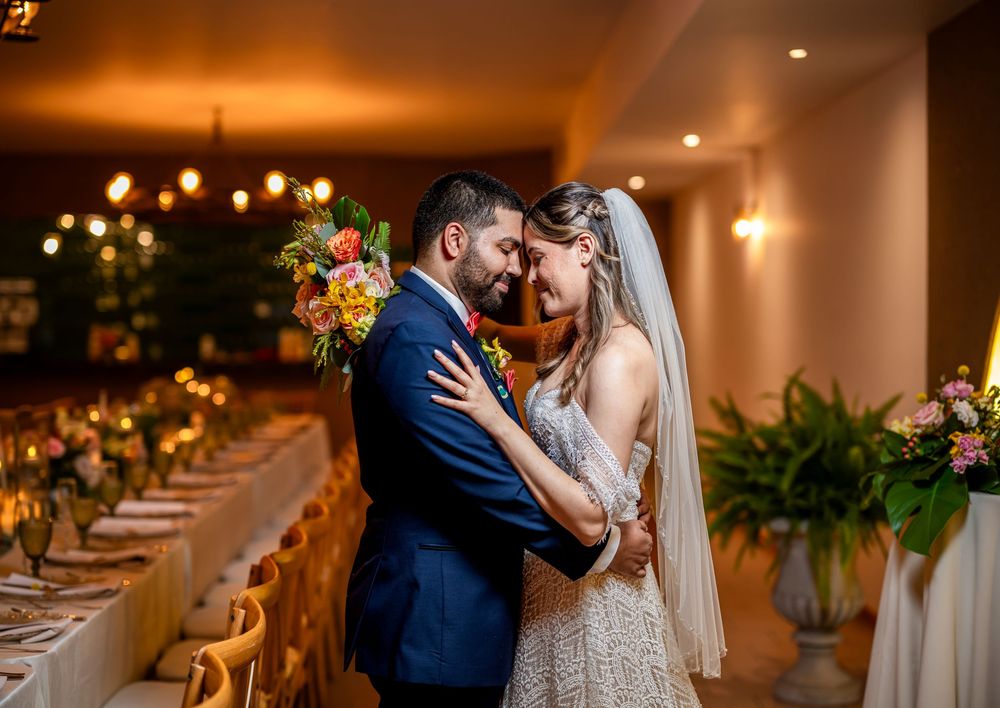 Newlywed couple shares intimate moments in warm, amber-lit wedding venue with decorative lighting and greenery.
