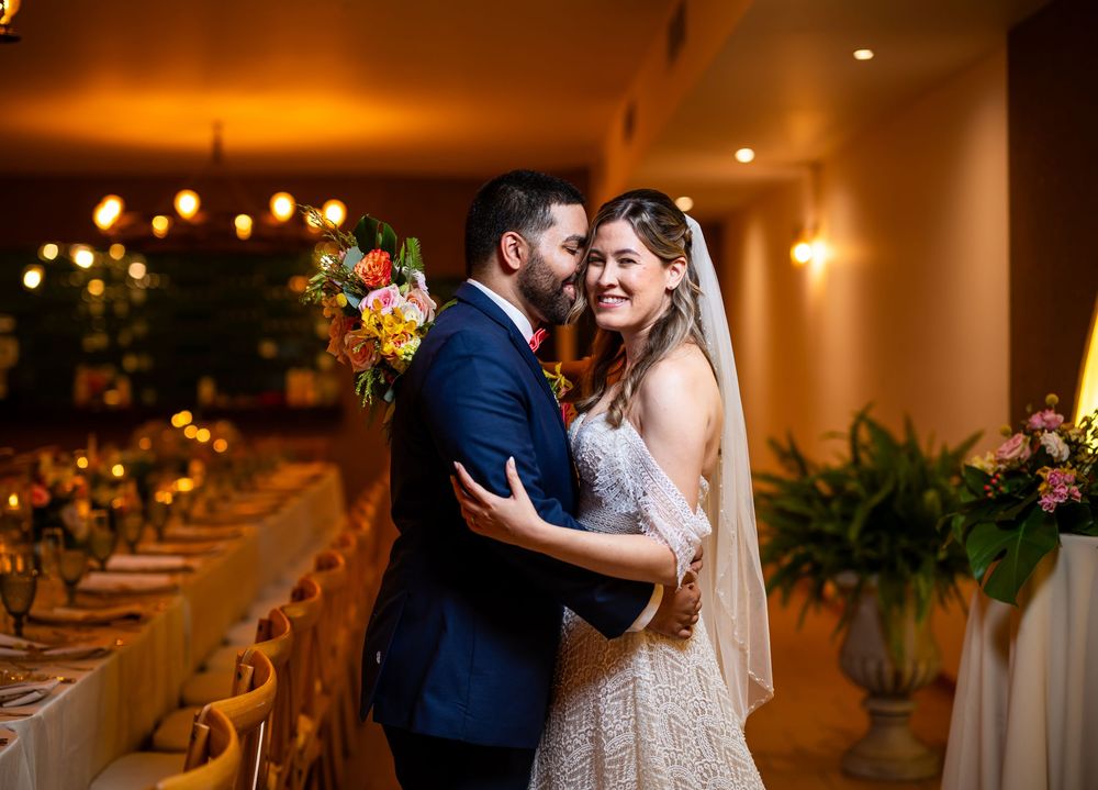 A newlywed couple shares an intimate moment in a warmly lit reception venue with floral decorations and long tables.