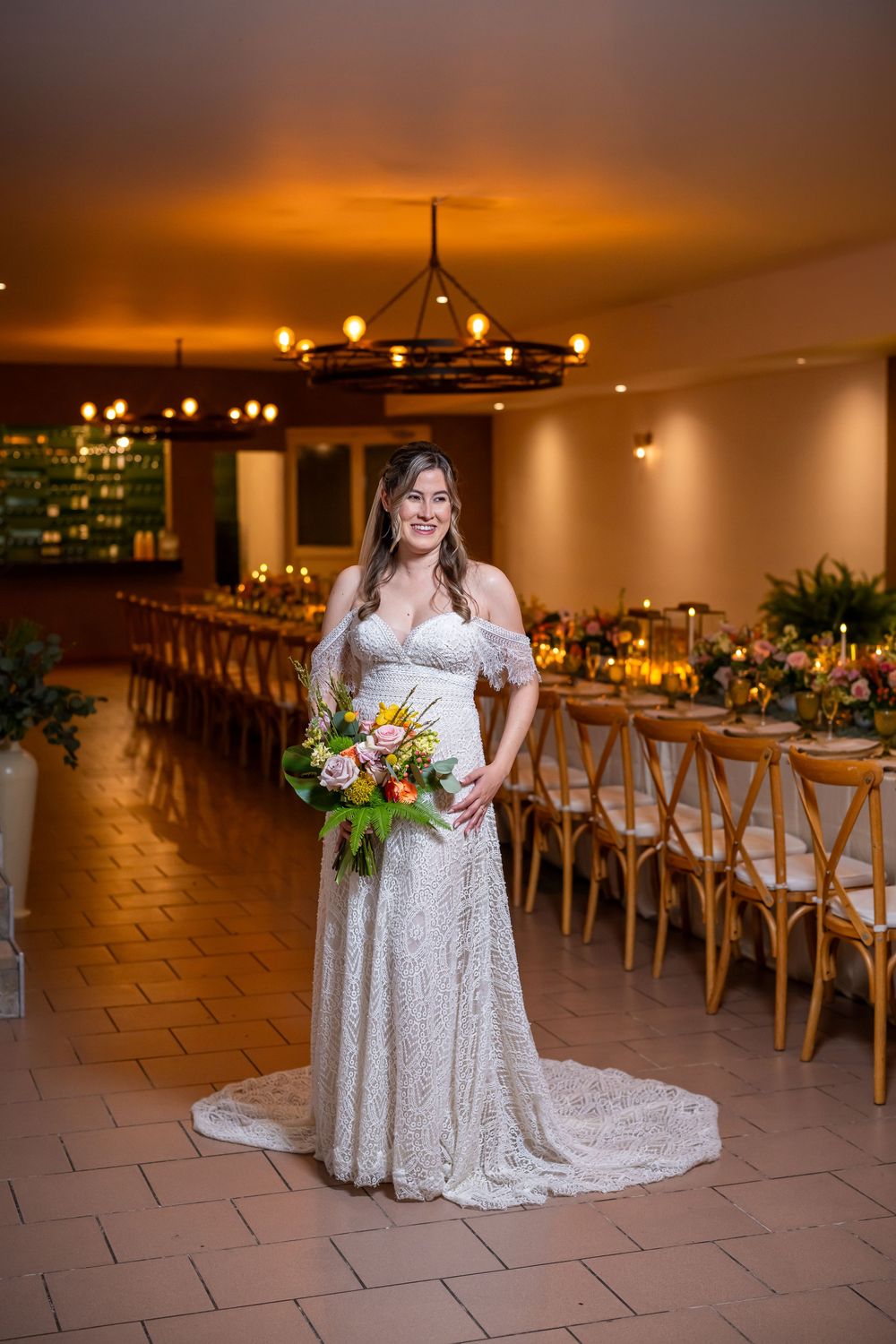 Elegant bride in lace off-shoulder gown holding vibrant bouquet in reception venue with rustic wooden tables and warm lighting.
