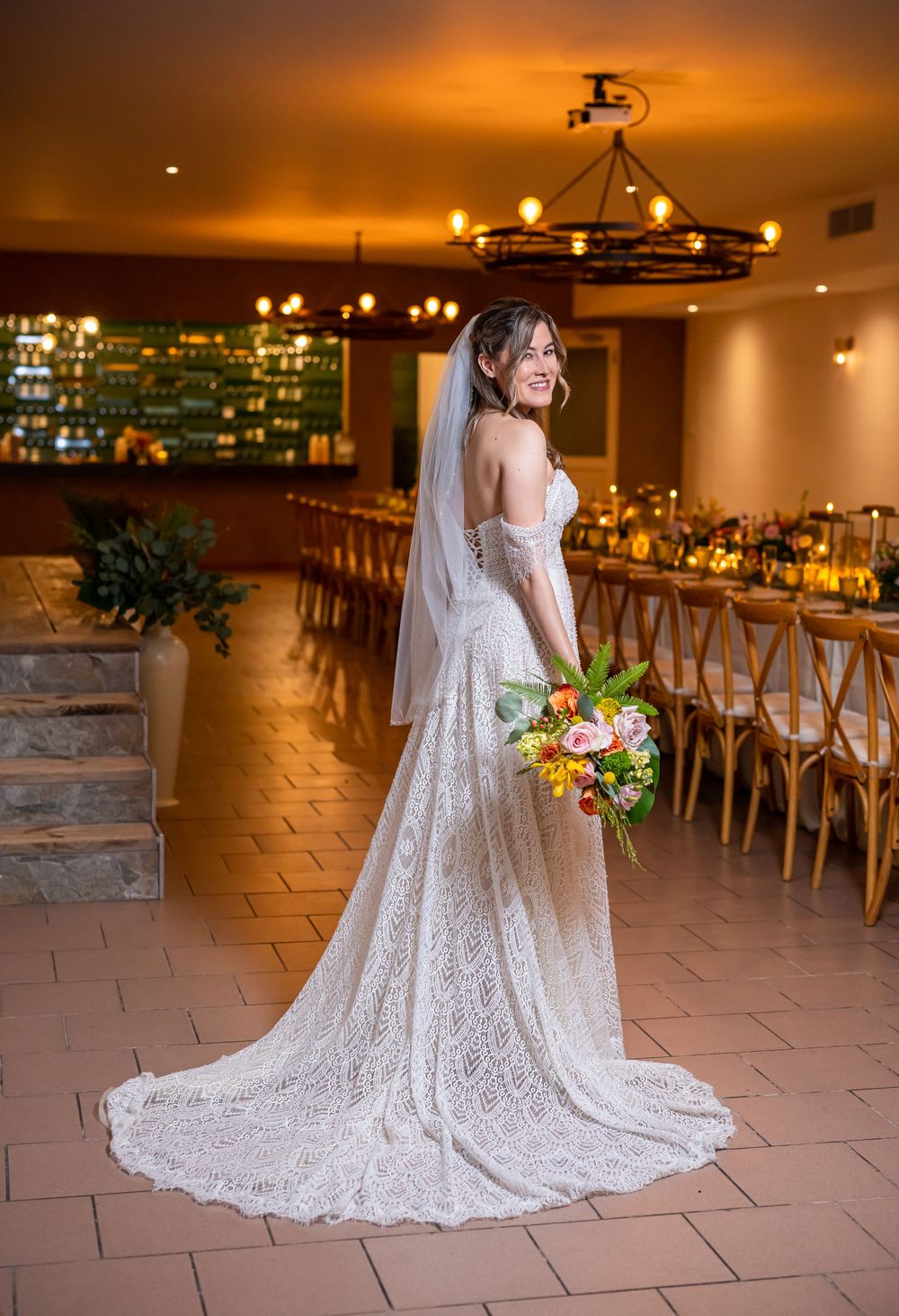 White lace wedding dress train spreads elegantly across stone floor in warmly lit rustic venue.