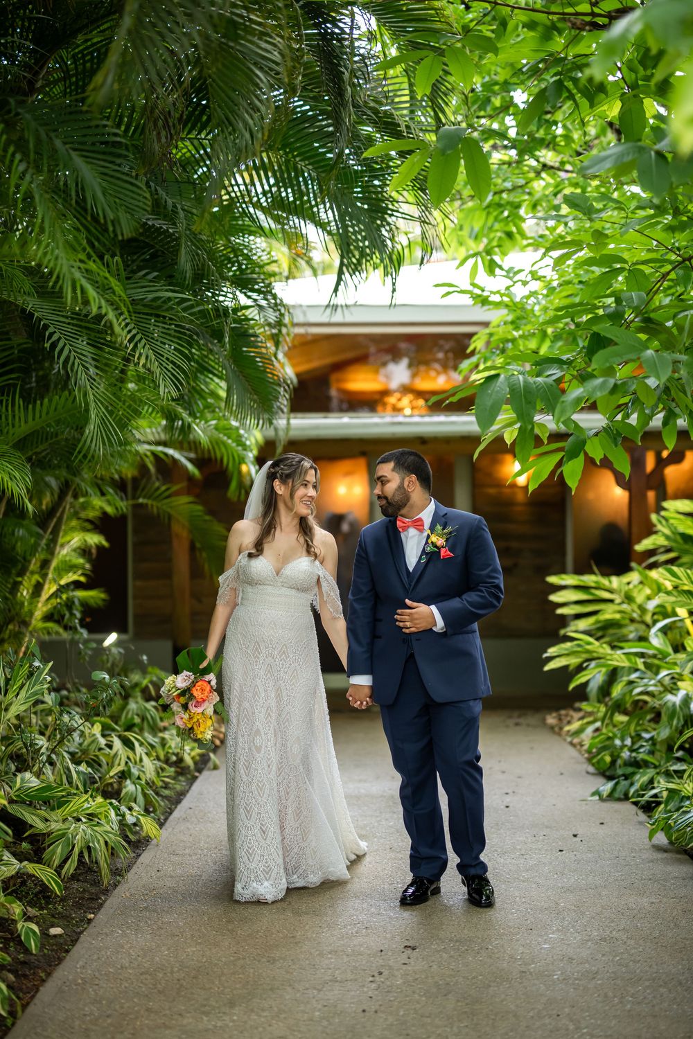 A romantic wedding couple walks along a lush garden path surrounded by tropical plants and trees.