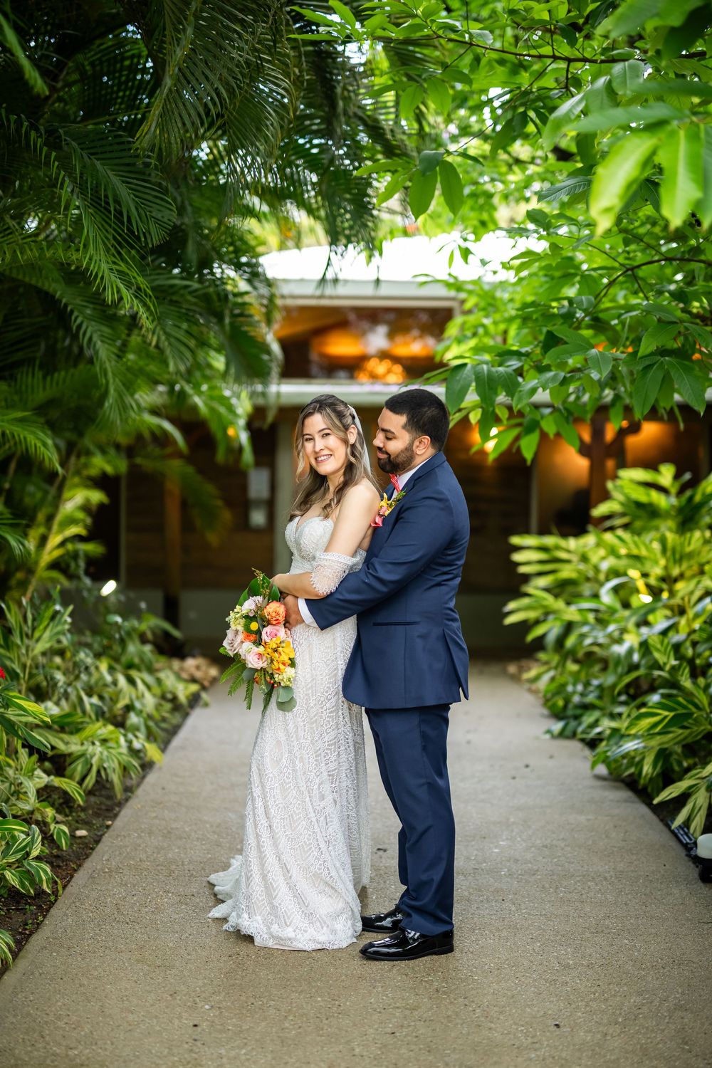 A couple shares a romantic moment on a garden path surrounded by lush tropical foliage with warm lighting in the background.