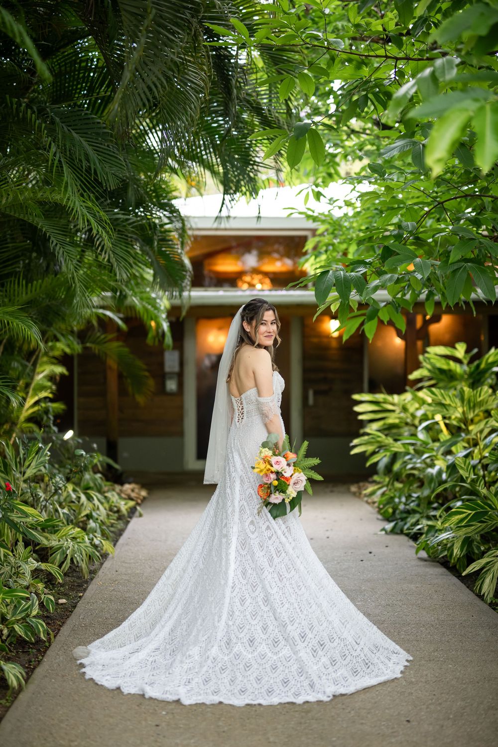 Elegant bridal portrait showing a flowing white lace wedding gown with cathedral train in a lush tropical garden setting.