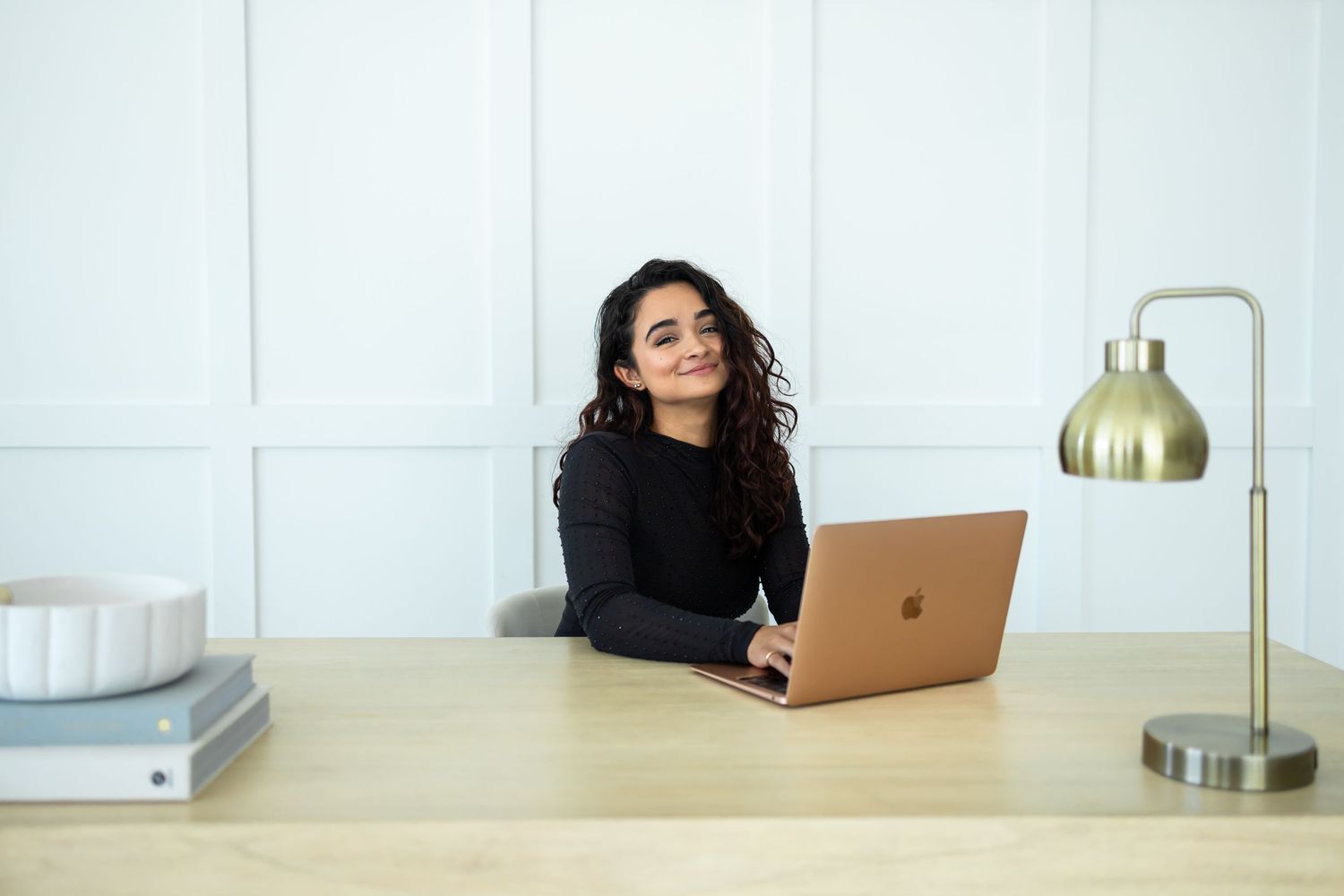 Professional working at wooden desk with MacBook laptop and brass desk lamp in bright modern office setting.