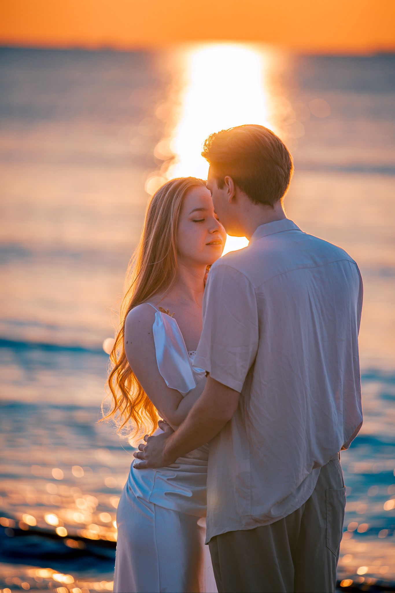 A couple in white outfits embraces on a beach at sunset with golden light reflecting off the ocean waves.