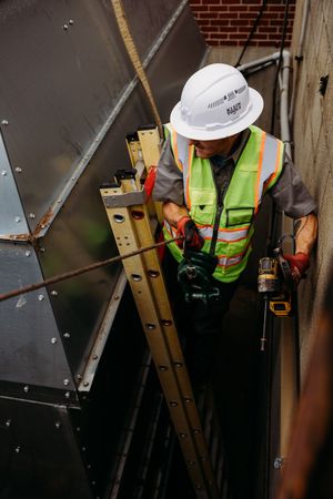 Construction worker in safety vest and hard hat carefully positions a ladder against a brick wall during repair work.