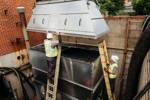 Workers inspect ventilation equipment on the roof of a brick commercial building.