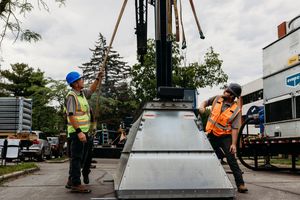 Two construction workers in safety vests and hard hats working on installing metal equipment with a crane outdoors.