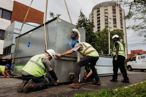 Workers in safety vests collaborate to move a large container on an urban street with buildings in the background.