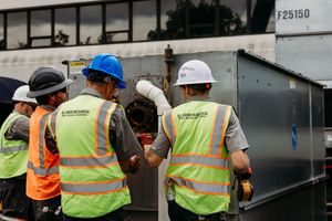Construction workers in safety vests and hard hats gather around equipment at a job site.