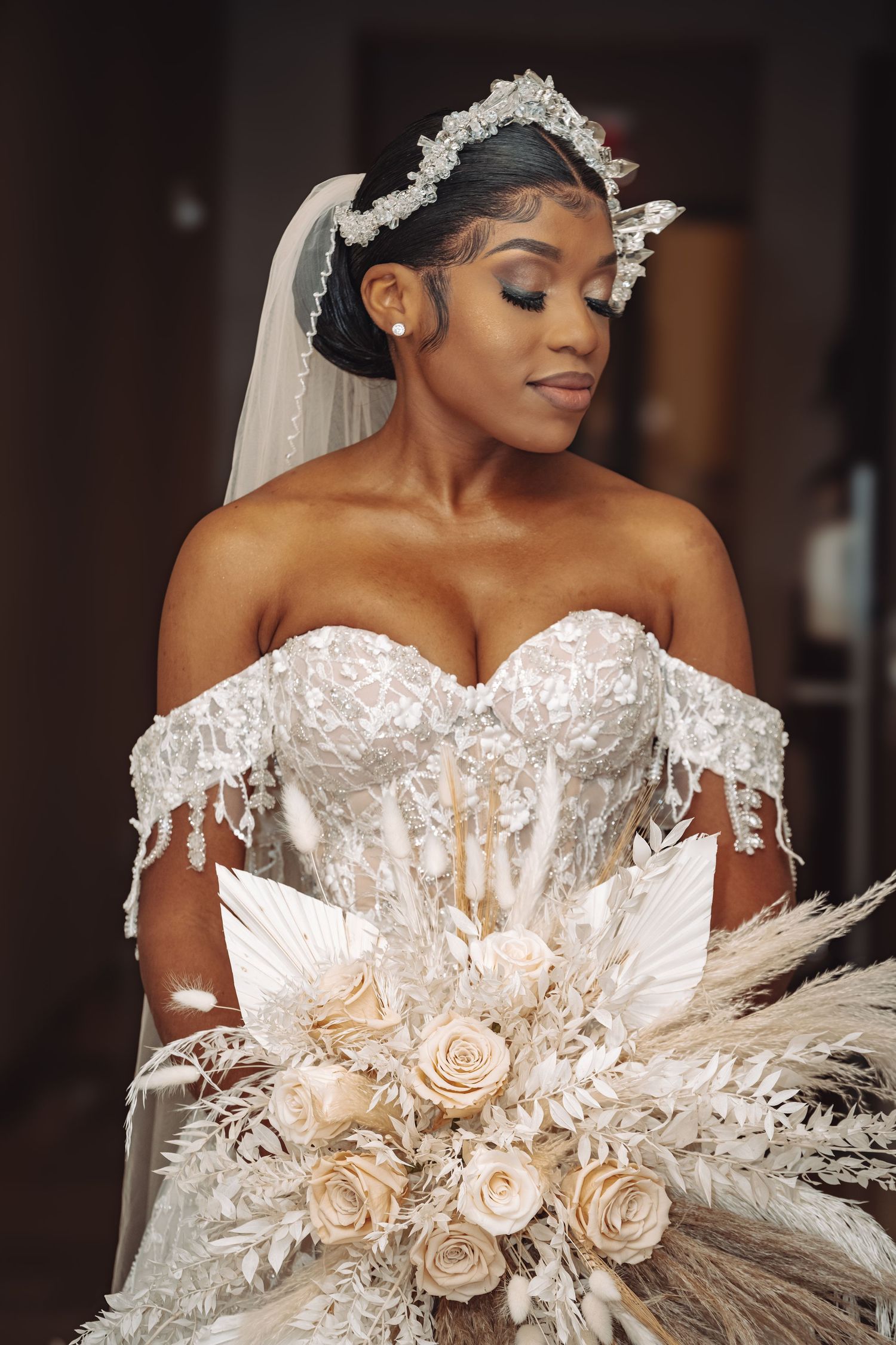 Beautiful bride wearing white off-shoulder wedding gown with lace details and holding a cream rose bouquet with pampas grass.