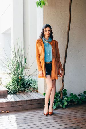 A woman poses on a wooden deck wearing a burnt orange cardigan, denim shirt, black shorts and brown heels.