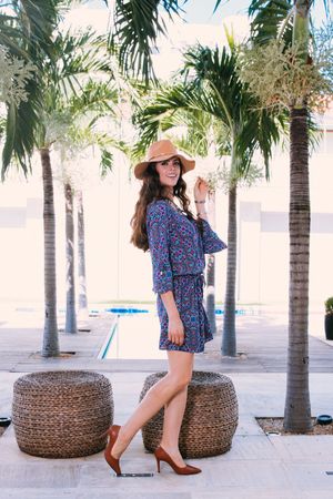 A woman in a floral romper and sunhat poses on a woven ottoman among palm trees in an outdoor courtyard.
