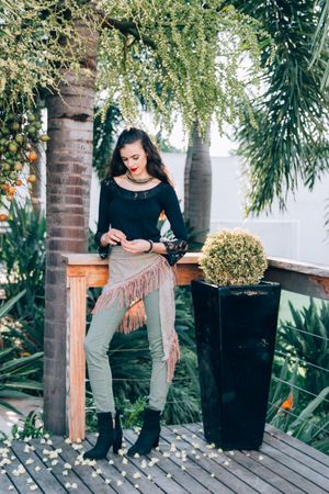 On a wooden deck with tropical plants, someone in black top and fringe pants leans against a pillar by a decorative planter.