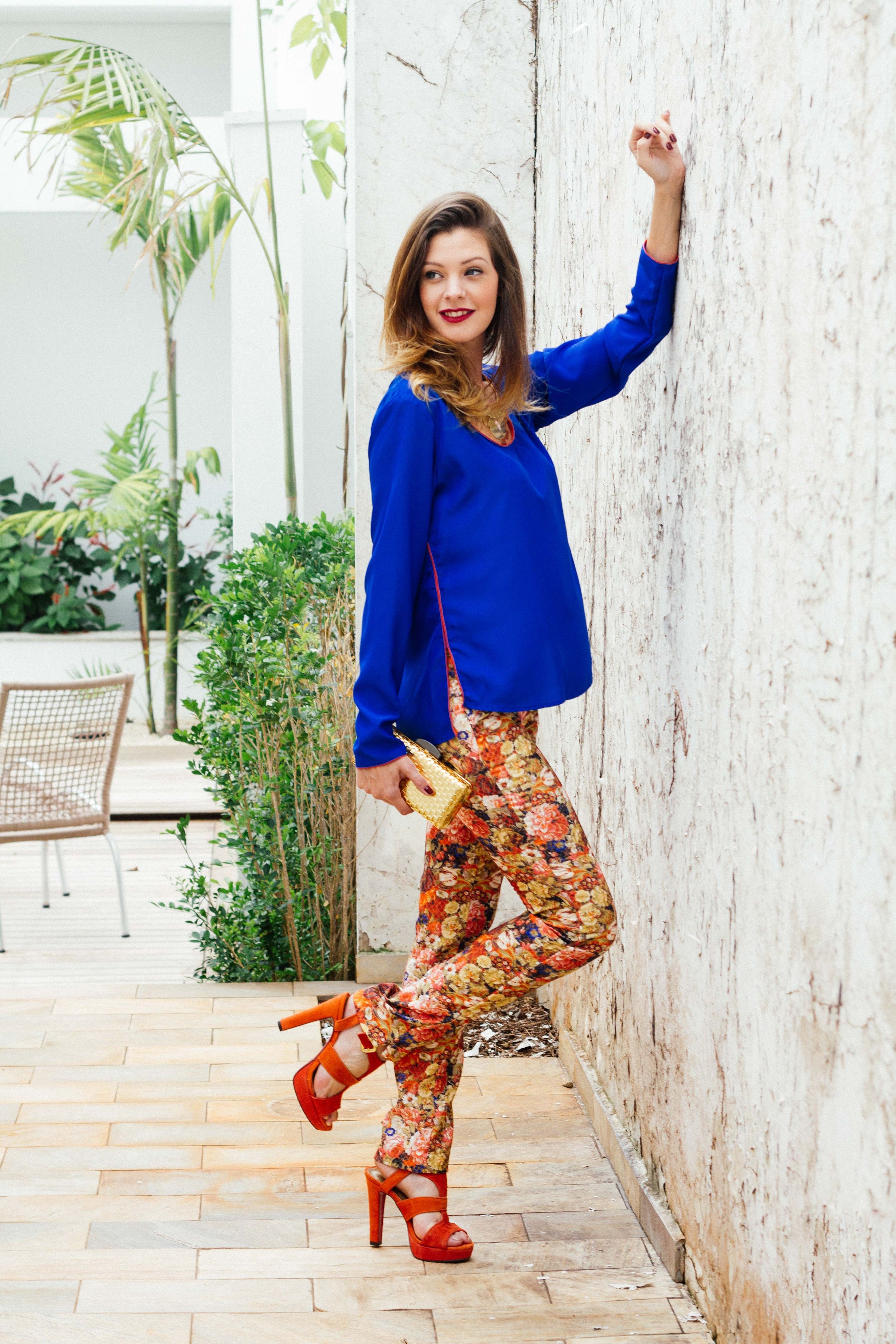 Striking fashion look with bright royal blue sweater paired with floral print pants and red platform sandals against white wall.